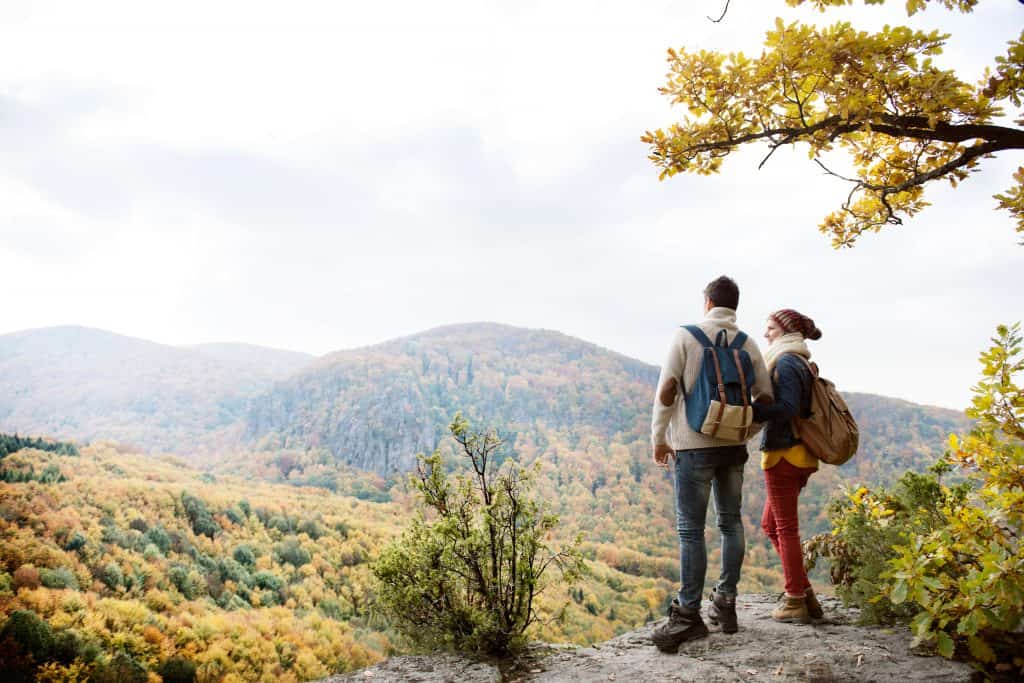 Beautiful couple with backpacks standing on a rock against colorful autumn forest, rear view