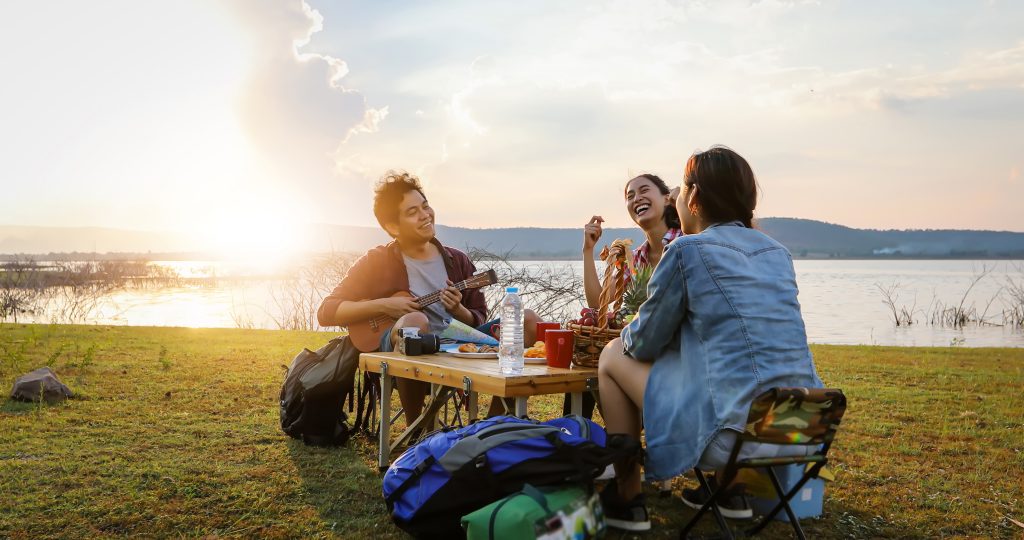 A group of Asian friends tourist drinking and playing guitar together with happiness in Summer while having camping near lake at sunset